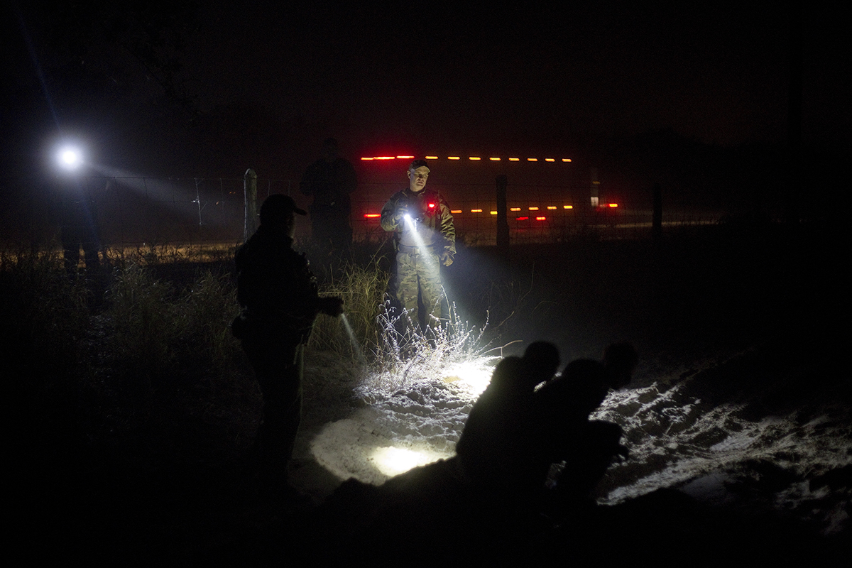 Border Patrol agent Christopher Hamer waits with apprehended migrants for transport vehicles on a ranch in Brooks County, Texas. Migrants walking here are avoiding the checkpoint in Falfurrias, Texas, along Rt. 281 North (background) to San Antonio in February 2015. Photo by Kirsten Luce.